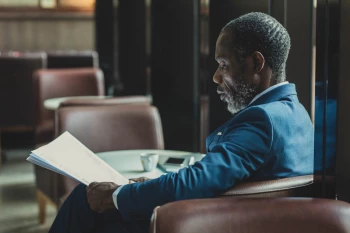 A man in a suit sitting in a chair, reading the news on a paper