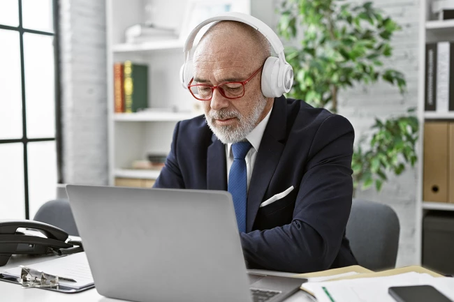 An older man in a suit wearing headphones, focused on working